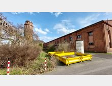 Industrielle Außenanlage mit einem roten Backsteingebäude im rechten Bildbereich und einem runden Wasserturm im Hintergrund. Im Vordergrund befinden sich Büsche, eine asphaltierte Fläche und zwei gelbe Metallcontainer.