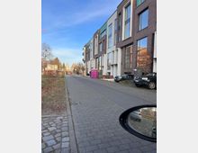 Straßenansicht eines modernen Wohnviertels mit mehrstöckigen Gebäuden aus Klinker und großen Fenstern rechts an der Straße. Links ist ein Grünstreifen mit kahlen Bäumen, im Vordergrund sind parkende Autos sowie ein auffälliges pinkes mobiles Toilettenhäuschen zu sehen.