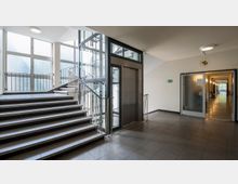 Treppenhaus mit modernem Design, bestehend aus einer Treppe mit Metallgeländer, einem verglasten Aufzug sowie großen Fenstern, die Tageslicht hereinlassen. Rechts führt ein Korridor mit Türen und Wandlampen in einen weiteren Bereich.