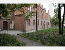 Backstein-Kirchgebäude mit gotischen Spitzbögen und schmalen, verzierten Strebepfeilern, umgeben von einem gepflasterten Weg und einem kleinen Garten mit Sträuchern und Bäumen. Im Hintergrund sind weitere Bäume und ein hoher, weißer Turm sichtbar.