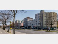 Straßenszene mit mehrstöckigen Wohn- und Geschäftsgebäuden, teils in farbigen Fassaden, sowie einer Bushaltestelle und parkenden Autos auf beiden Straßenseiten. Bäume ohne Laub säumen den Gehweg, und der Himmel ist klar.