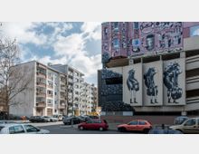 Ein urbaner Straßenabschnitt mit parkenden Autos und mehreren Wohngebäuden. Ein markantes Gebäude rechts zeigt großflächige Wandmalereien mit surrealen Motiven und technischen Elementen, während links ein schlichtes Wohnhaus mit Balkonen zu sehen ist.