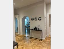 Ein heller Eingangsbereich mit hellgrauen Wänden und einem Fischgrätparkettboden. An der Wand hängt eine Reihe dekorativer Teller mit Gesichtern, darunter steht ein schwarzes, minimalistisches Konsolentischchen mit einer schwarzen Vase und einem Raumduftbehälter. Im Hintergrund führt ein Rundbogen in einen weiteren Raum mit Sitzmöbeln.