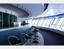 Innenraum einer modernen Bar in einem Aussichtsturm mit geschwungener Theke, Barhockern mit schwarzen Sitzen und großen, schrägen Fenstern, die viel Licht hereinlassen. Die Decke ist abgerundet, und die Einrichtung wirkt futuristisch mit klaren Linien und kühlen Farbtönen.