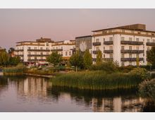 Großes, modernes Gebäude mit hellen Fassaden und mehreren Balkonen, gelegen an einem See mit Schilfbewuchs im Vordergrund. Im Umfeld des Gebäudes befinden sich Grünflächen, Bäume und ein kleiner Bereich mit Sitzmöglichkeiten.