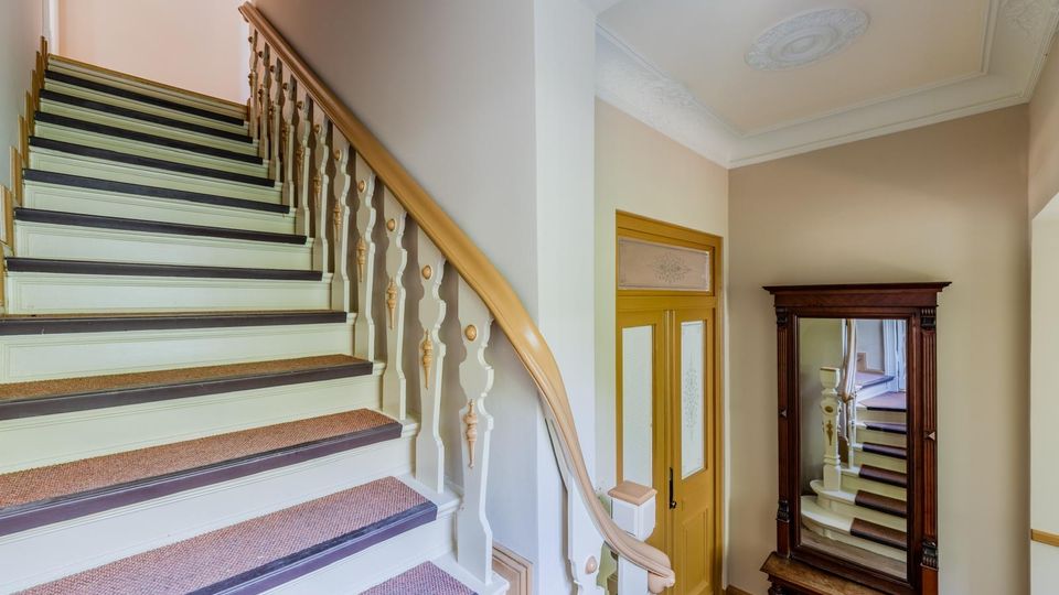 Innenansicht eines Altbau-Treppenhauses mit breiter Holztreppe, hell gestrichenen Stufen, Teppichläufer und geschwungenem Geländer mit Zierelementen. Daneben stehen eine gelbe Innentür mit Glasfeld und ein großer, dunkel gerahmter Spiegel; die Decke hat Stuckornamente.