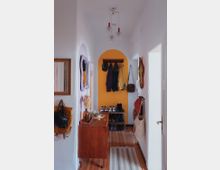 Schmaler Flur mit Holzboden, einer gelben Akzentwand mit Haken für Jacken und Hüte sowie einem Schuhregal darunter. Links steht eine kleine Kommode mit Dekorationen, und rechts hängen Taschen an der Wand.