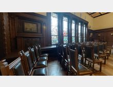 Ein historischer Raum mit Holzwänden und einer Reihe von hölzernen Stühlen, die mit schwarzem Leder gepolstert sind. Große Buntglasfenster mit detailreichen Motiven dominieren die Wand und lassen farbiges Licht in den Raum fallen.