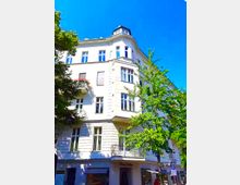 Mehrstöckiges, hell verputztes Eckwohnhaus im Gründerzeitstil mit hohen Fenstern, Erkern und einem kleinen Balkon, aufgenommen aus Straßenperspektive. Im Vordergrund stehen grüne Straßenbäume, darüber ein klarer blauer Himmel.