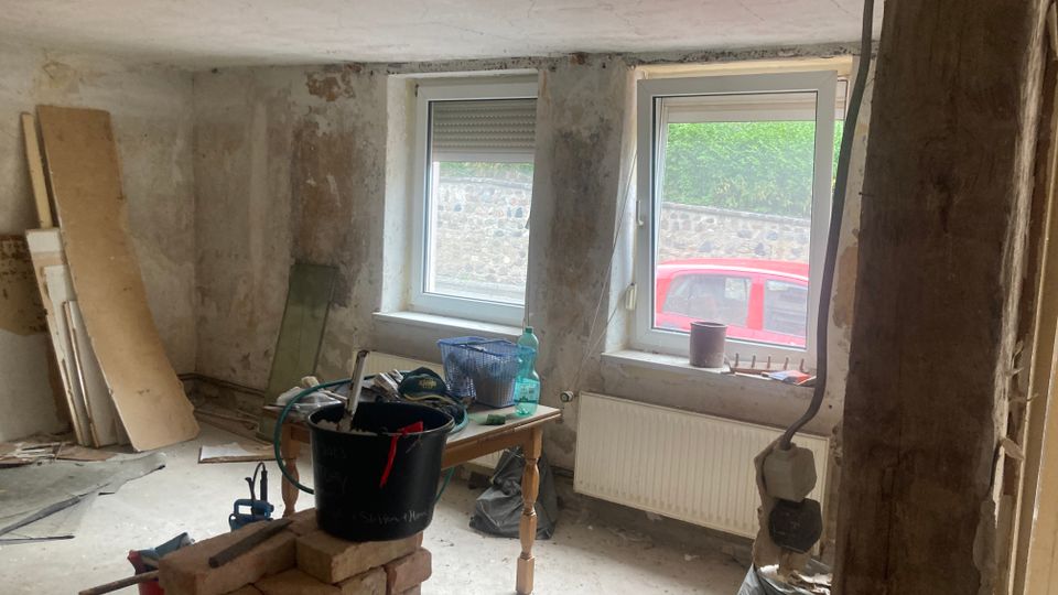 Ein unfertiger Innenraum mit verputzten, teils beschädigten Wänden, zwei Fenstern mit Blick nach draußen, und diversen Werkzeugen und Baumaterialien auf dem Boden sowie einem Tisch. Im Vordergrund steht ein kleiner Stapel Ziegelsteine und ein schwarzer Eimer.