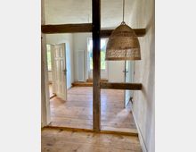 Ein helles Zimmer mit Holzboden und weiß verputzten Wänden. Sichtbare Holzbalken trennen den Raum, im Vordergrund hängt eine Lampe mit einem Schirm aus geflochtenem Material; im Hintergrund sind große Fenster und geöffnete Türen sichtbar.