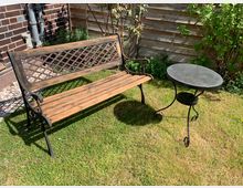 Holzbank mit Metallgestell und dekorativer Rückenlehne neben einem runden Metalltisch auf einer Rasenfläche, im Hintergrund ein Holzzaun, Zierpflanzen und eine Ziegelhauswand.