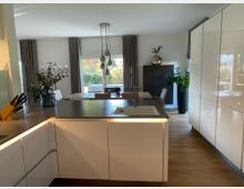 Moderne, offene Küche mit weißen Hochglanzschränken, einer zentralen Kücheninsel mit Spüle, Holzfußboden und großen Fenstern, die viel Tageslicht hineinlassen. Im Hintergrund befindet sich ein Essbereich mit Tisch, Stühlen und großen Zimmerpflanzen.