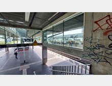 Ein moderner S-Bahnhof mit überdachten Bahnsteigen, Glaswänden und klar markierten Gleisen. Graffiti bedeckt die Betonwände, und eine Treppe führt zu den Bahnsteigen, auf denen Reisende warten.