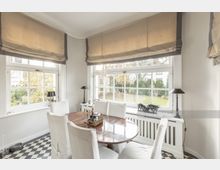 Esszimmer mit großem Holztisch und vier weißen Stühlen, bodentiefen Sprossenfenstern mit Blick in einen Garten sowie schwarz-weißen Schachbrettfliesen. Im Raum befinden sich helle Rollläden, dezente Stehlampen und Dekorationselemente.