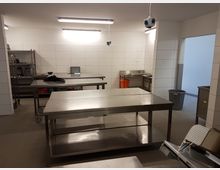 Großer, heller Arbeitsraum mit weißen, gefliesten Wänden und grauem Boden. Im Raum befinden sich mehrere Edelstahl-Arbeitstische, ein Regal mit Küchenutensilien, ein Waschbecken sowie Mülleimer und orangefarbene Behälter.