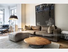 Modernes Wohnzimmer mit einer braunen Eckcouch, einem passenden Hocker und einem runden Holztisch auf einem grob gewebten Teppich. Im Hintergrund sind große Fenster mit viel Tageslicht, ein schwarzer Ledersessel mit Hocker sowie ein abstraktes Gemälde an der Wand zu sehen.