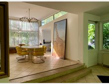 Essbereich mit gelben Retro-Stühlen um einen runden Tisch, großen vertikalen Lamellenfenstern, die Blick ins Grüne bieten, sowie einer auffälligen Giraffen-Abbildung in einem Rahmen an der Wand. Der Boden ist mit beigefarbenen Fliesen gestaltet, und im Vordergrund führen Stufen in den Raum.