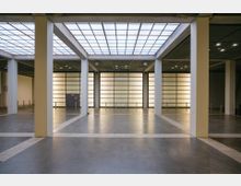 Weitläufiger Innenraum mit hellen, beleuchteten Wänden, Glasdach und symmetrischen Säulen, die eine offene und minimalistische Architektur zeigen. Der Boden ist aus poliertem Beton, mit weißen Markierungen, die den Raum unterteilen.