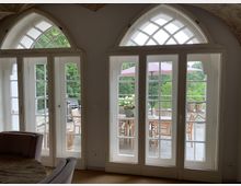 Große weiße Doppelflügeltüren mit gotischen Bögen führen zu einer Terrasse mit Holztisch, Stühlen, Pflanzen und einem Sonnenschirm. Im Hintergrund ist eine grüne Gartenlandschaft zu sehen.