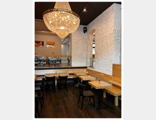 Innenansicht eines modernen Cafés mit weißen Backsteinwänden, einem dunklen Holzboden und Holzmöbeln. Im Mittelpunkt hängt ein großer Kronleuchter, und kleine Tische sind mit frischen Blumen dekoriert.