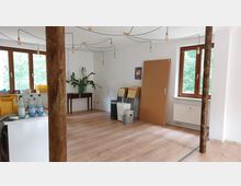 Ein heller Raum mit Holzboden, weißen Wänden und mehreren großen Fenstern mit Holzrahmen, die Ausblick ins Grüne bieten. Im Raum befinden sich gelagerte Stühle, ein kleiner Tisch mit einer Pflanze, Mülleimer und diverse Gegenstände wie Wasserflaschen und gelbe Kunststoffkisten; von der Decke hängen mehrere Glühbirnen an schwarzen Kabeln herunter.