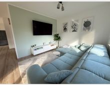 Wohnzimmer mit heller Einrichtung, bestehend aus einer großen hellblauen Polsterwohnlandschaft, einem weißen TV-Unterschrank mit Dekoration, einem an der Wand montierten Flachbildfernseher, drei abstrakten Pflanzenbildern an der Wand und einem kleinen Beistelltisch. Der Raum hat eine mintgrüne Akzentwand, Holzboden und wird durch ein großes Fenster auf der rechten Seite hell beleuchtet.