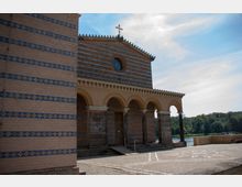 Gelb-braune Backsteinkirche mit blauen Ziermustern und einer Fassade mit Arkadenbögen, darüber ein Giebeldach mit einem Kreuz. Die Kirche steht in einer offenen Umgebung mit Blick auf einen Fluss und bewaldete Ufer im Hintergrund.