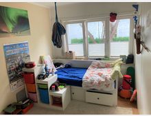 Ein Kinderzimmer mit Einzelbett und blauer Bettwäsche, daneben ein Regal mit bunten Aufbewahrungsboxen und Spielsachen. An der Wand hängen ein Poster mit Nordlichtern, ein Supercross-Poster und diverse Gegenstände; das Zimmer hat große Fenster mit Blick auf Bäume und Rasen.