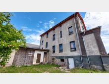 Ein verwittertes mehrstöckiges Gebäude mit grauer Fassade, mehreren Fenstern und rostigen Regenrinnen, flankiert von einem angebauten Schuppen mit Holztoren. Im Vordergrund ist ein grüner Baum und eine kleine Wiese zu sehen, darüber ein blauer Himmel mit einigen Wolken.