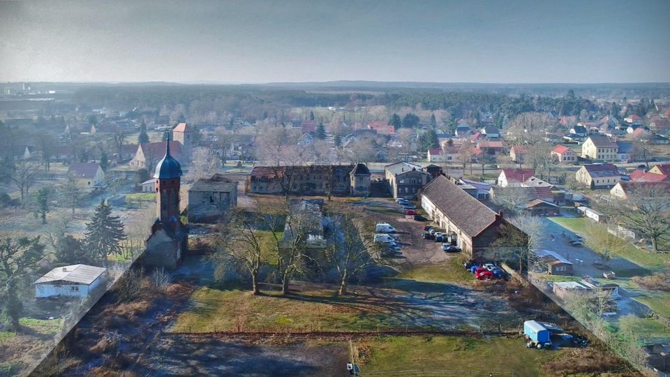 Luftaufnahme eines dörflichen Areals mit einem historischen Kirchen- oder Turmgebäude mit dunkler Kuppel, umgeben von Wiesen, Bäumen und mehreren länglichen Wirtschaftsgebäuden. Im Hintergrund erstreckt sich ein Wohngebiet mit Einfamilienhäusern und Feldern bis zum Horizont.