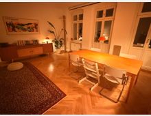 Gemütlicher Essbereich in einer Wohnung mit großem Holztisch, weißen Stühlen und orangefarbenen Pendelleuchten über dem Tisch. Helle Wände, Parkettboden im Fischgrätmuster, großer Teppich, Sideboard mit Büchern, Zimmerpflanze sowie mehrere hohe Fenster und eine Balkontür prägen den Raum.