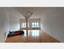 Weitläufiger, leerer Raum mit hellen Wänden, einem Holzboden und drei großen Fenstern, die natürliches Licht hereinlassen. Im Vordergrund befinden sich eine aufblasbare Matratze, eine Decke und ein Stuhl.