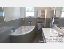 Modernes Badezimmer mit einer Eckbadewanne, grauen Fliesen und einer weißen Wand. Zu den Ausstattungselementen gehören ein beleuchteter Spiegel, ein Waschbecken, ein Heizkörper mit Handtüchern und ein Fenster mit natürlichem Lichteinfall.