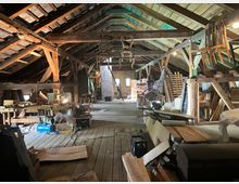 Ein geräumiger Dachboden mit freiliegenden Holzstützen und -balken sowie einer schrägen Holzdecke. Der Raum ist mit Holzplanken und handwerklichen Materialien gefüllt, darunter Werkbänke, Werkzeuge, Holzstapel und gelagerte Gegenstände.