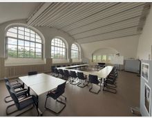 Ein heller Seminarraum mit hohen Decken, mehreren großen, bogenförmigen Fenstern und einer U-förmigen Anordnung von Tischen mit dunkel gepolsterten Stühlen. An der Wand befinden sich Whiteboards und eine Pinnwand, der Boden ist mit einem glatten, beigen Belag ausgestattet.