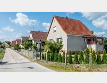 Reihen von Einfamilienhäusern mit roten Ziegeldächern entlang einer gepflasterten Straße, mit Vorgärten und kleinen Hecken hinter Zäunen; im Hintergrund blauer Himmel mit vereinzelten Wolken.