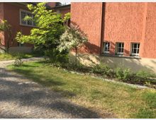 Roter Gebäudewand mit mehreren kleinen weißen Fenstern, umgeben von einem grünen Gartenbereich mit Bäumen, Sträuchern und Rasenflächen. Vor dem Gebäude führt ein gepflasterter Weg entlang.