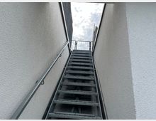 Treppenaufgang aus Metall mit Geländern auf beiden Seiten, zwischen weißen Putzwänden eingezäunt und oben mit einer metallenen Tür abgeschlossen; im Hintergrund ist der Himmel mit Wolken sichtbar.