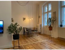 Ein Wohnzimmer mit Holzdielenboden, weißen Wänden und großen Fenstern mit Rundbögen, die viel Licht hereinlassen. Im Raum befinden sich ein Esstisch mit gemischten Stühlen, hängende Glühbirnen als Beleuchtung sowie mehrere Zimmerpflanzen in Töpfen.