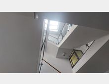 Ansicht eines modernen Treppenhauses mit klaren Linien, weißen Wänden und einem Geländer aus Glas und Metall. Licht fällt durch ein großes Fenster im oberen Bereich, das mehrere Etagen verbindet.