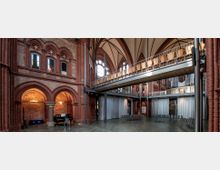 Innenansicht einer umgebauten Kirche mit rotem Backsteingemäuer und spitzbogiger Decke. Der Raum verfügt über eine moderne Zwischengalerie mit Sitzplätzen, große Fenster mit historischen Bögen und Vorhänge, die flexible Raumtrennungen ermöglichen.