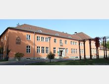 Ein zweistöckiges Gebäude mit orangefarbener Fassade und vielen weißen Fenstern, in schlichtem symmetrischen Stil. Vor dem Gebäude befindet sich ein gepflegter Gehweg, eine Rasenfläche und eine überdachte Pergola-Struktur.