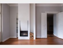 Heller, leerer Wohnraum mit hohen, hellgrauen Wänden und Holzdielenboden, in der Mitte ein weißer Kamin mit gläsernem Einsatz und daneben gestapeltem Brennholz. Rechts führen geöffnete, weiße Doppeltüren in einen angrenzenden Raum; links sind schmale, eingebaute Regale zu sehen.