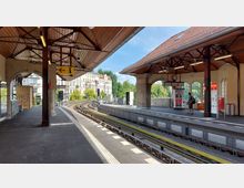 Ein oberirdischer Bahnhof mit zwei Bahnsteigen und einem geschwungenen Gleis. Die Überdachung besteht aus Holz und Stahlträgern, mit großen Fenstern in den Hintergrundwänden, umgeben von grünen Bäumen und Wohngebäuden.
