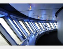 Innenraum einer Aussichtsplattform mit modernen Paneelwänden und schrägen Fenstern, durch die helles Licht einfällt. Ein geschwungener Handlauf führt entlang der Fensterfront, und blaue Beleuchtungselemente betonen die futuristische Gestaltung.