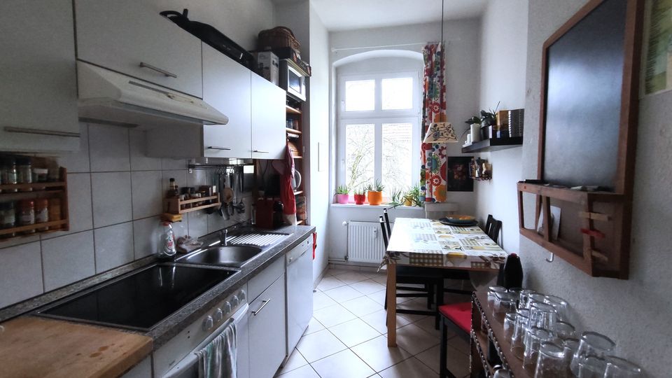 Narrow galley kitchen with white cabinets and tiled backsplash along one wall, a long countertop with sink and stove, and a small dining table with chairs on the opposite side. A tall arched window at the end lets in daylight, with potted plants on the sill, patterned curtains, wall shelves, and a chalkboard.