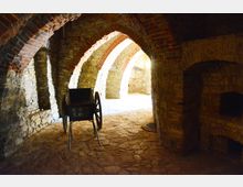 Gewölbekeller mit gemauerten Stein- und Ziegelbögen, unebenem Steinboden und schwachem Lichteinfall. Im Vordergrund steht eine historische, zweirädrige Holzkarre.