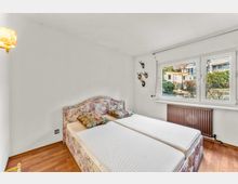 Schlafzimmer mit Doppelbett und gepolstertem, gemustertem Kopfteil auf Holzboden, weißen Wänden und Deckenleuchte. Großes Fenster mit Blick in einen begrünten Innenhof; darunter ein Heizkörper.