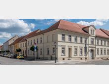 Straßenszene mit einem hellen, historischen Wohngebäude in einem Stadtviertel, gekennzeichnet durch rote Ziegeldächer, große Fenster und dekorative Fassaden. Am Straßenrand stehen geparkte Autos und ein Schild weist auf Parkmöglichkeiten hin.